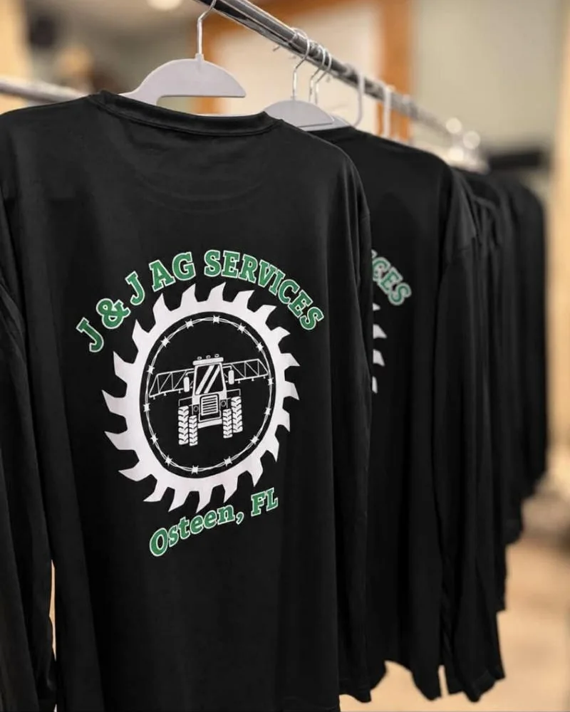 Black long-sleeve shirts on hangers printed with the J&J Ag Services saw-blade logo